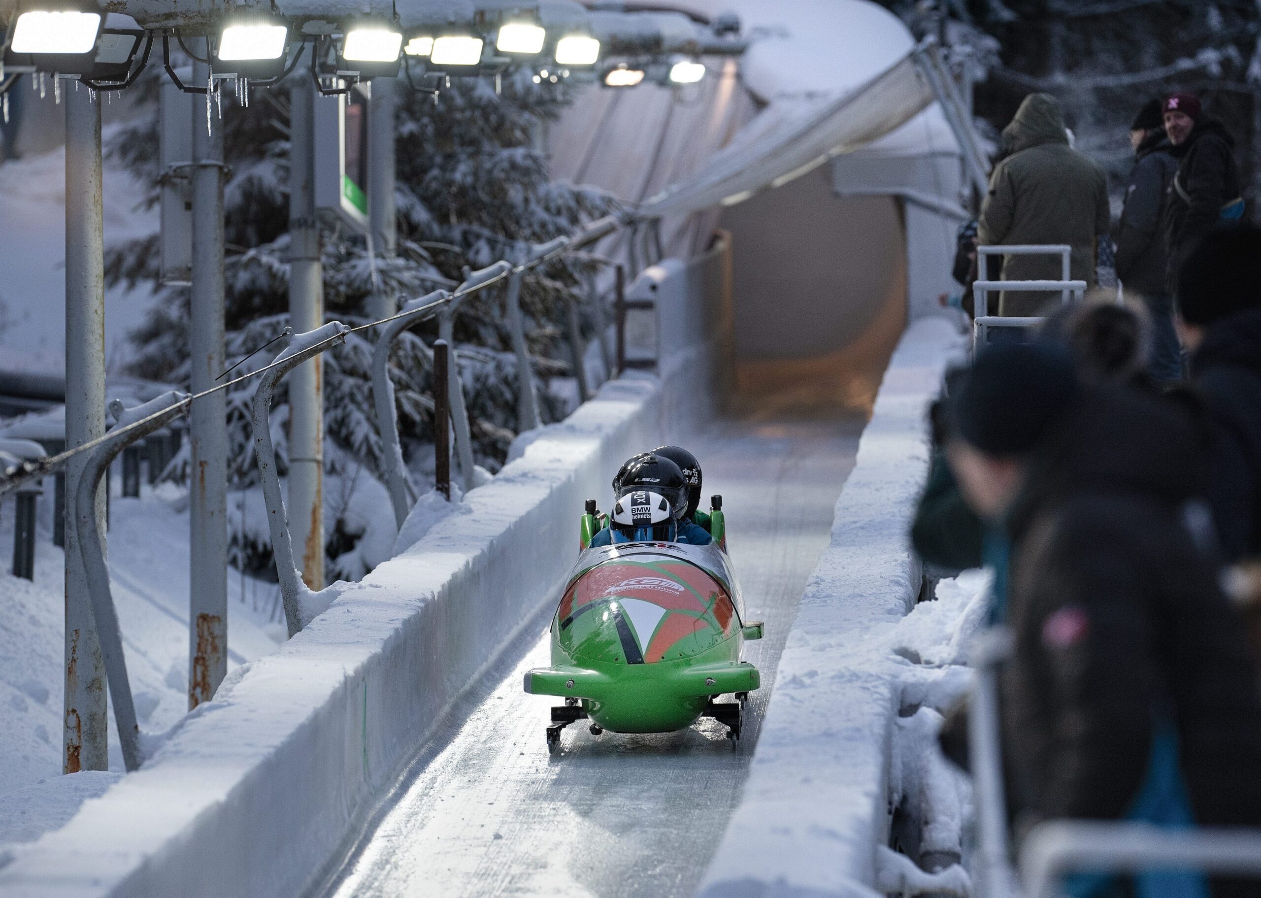 Ein grüner Bob mit zwei Athleten rast die vereiste Bobbahn hinunter, während Zuschauer in Winterkleidung zuschauen. Helle Scheinwerfer beleuchten die frostige Szenerie und machen sie selbst für ein Publikum der „Kinder-Edition“ spannend.