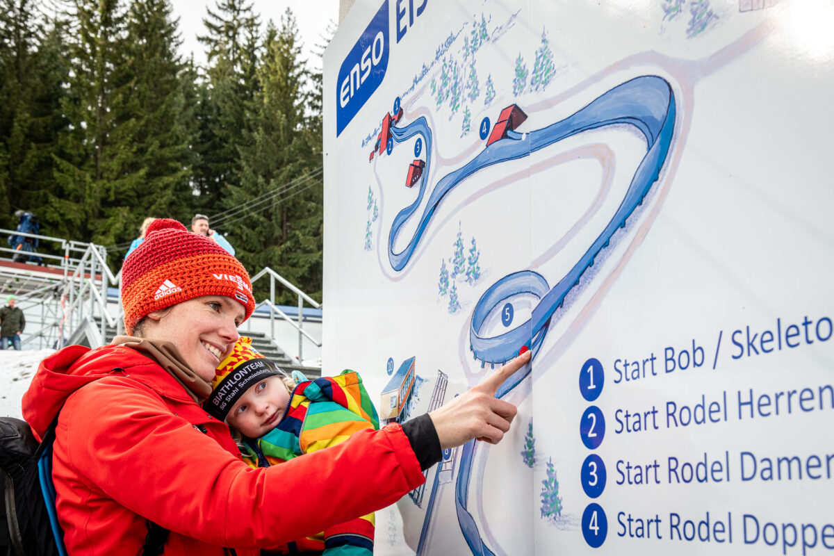 Ein lächelnder Erwachsener in roter Jacke und Mütze hält ein warm eingepacktes Kind im Arm und zeigt an einem verschneiten Tag im Freien auf den Streckenplan der Bobbahnführung Altenberg. Auf dem Schild sind die verschiedenen Startmöglichkeiten für die Rodelbahn aufgeführt, im Hintergrund sind Bäume und Tribünen zu sehen.