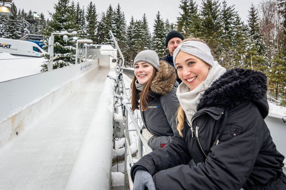Drei Menschen in Winterkleidung lächeln in der Nähe der Bobbahn in Altenberg, umgeben von verschneiten Bäumen und unter bewölktem Himmel, und genießen an einem schönen Wintertag im Freien eine Bobbahnführung.